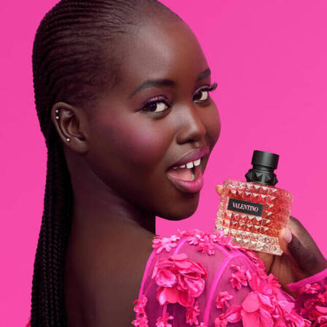 Eine lachelnde Frau mit dunkler Haut tragt ein pinkes Outfit mit Blumendetails und halt ein Parfumflakon vor einem pinken Hintergrund.
