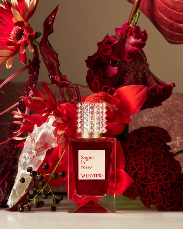 Ein rotes Parfumflaschchen umgeben von prachtvollen roten Blumen und Pflanzen, die eine elegante und sinnliche Atmosphare schaffen.