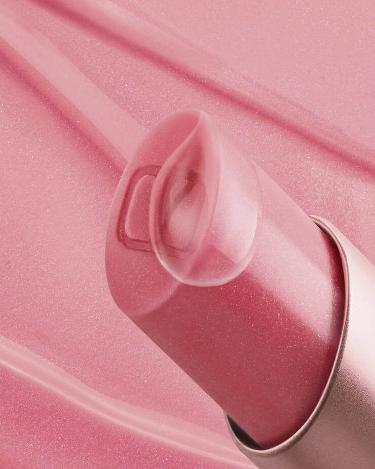 Eine Nahaufnahme eines pinkfarbenen Lippenbalsams mit seiner klaren, durchsichtigen Kappe, die leicht geoffnet ist und ein dezentes „V“-Logo im Inneren zeigt, alles vor einem zarten rosa Hintergrund mit schwachen Produktspuren.