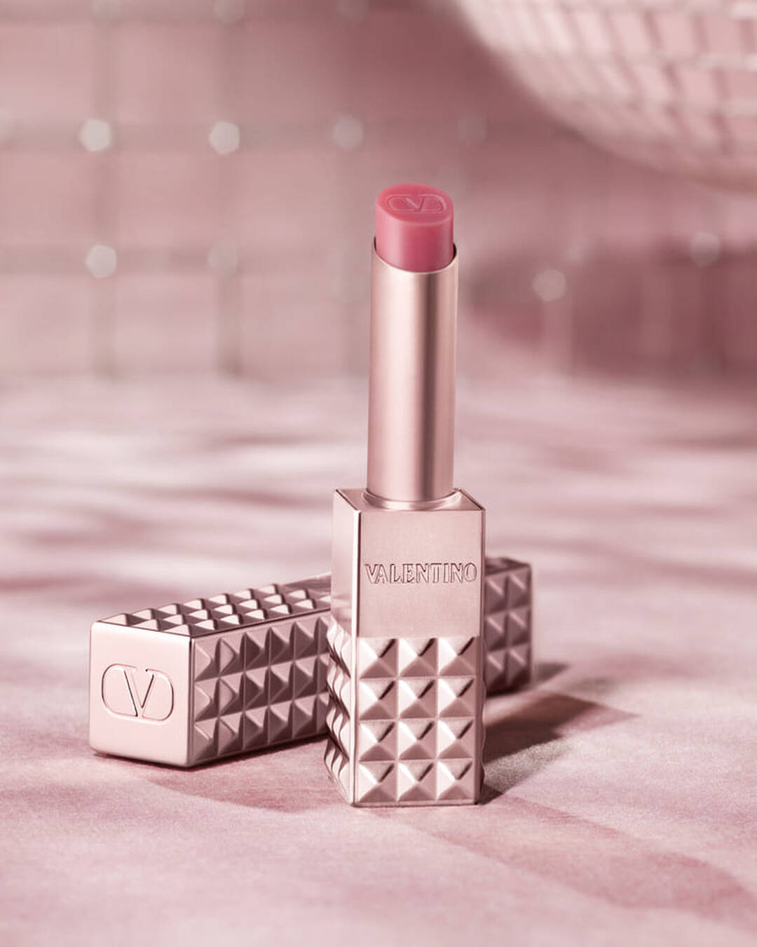 Ein rosegoldfarbenes Valentino Spike Lippenprodukt mit pinkfarbener Spitze steht aufrecht, daneben liegt sein passendes genietetes Etui. Beide Gegenstande sind auf einem weichen, glitzernden rosa Hintergrund platziert, der ein Gefuhl von Glamour vermittelt.