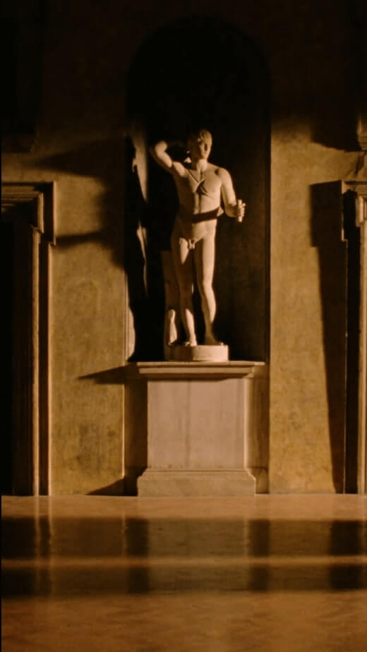 A classical statue of a man holding a cup, illuminated in a dimly lit alcove with textured walls.