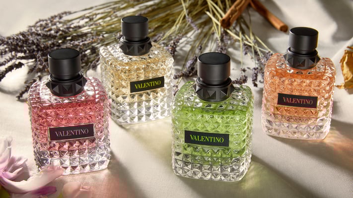 Vier Valentino-Parfümflakons in verschiedenen Farben stehen auf einem Tisch, umgeben von getrockneten Blumen.